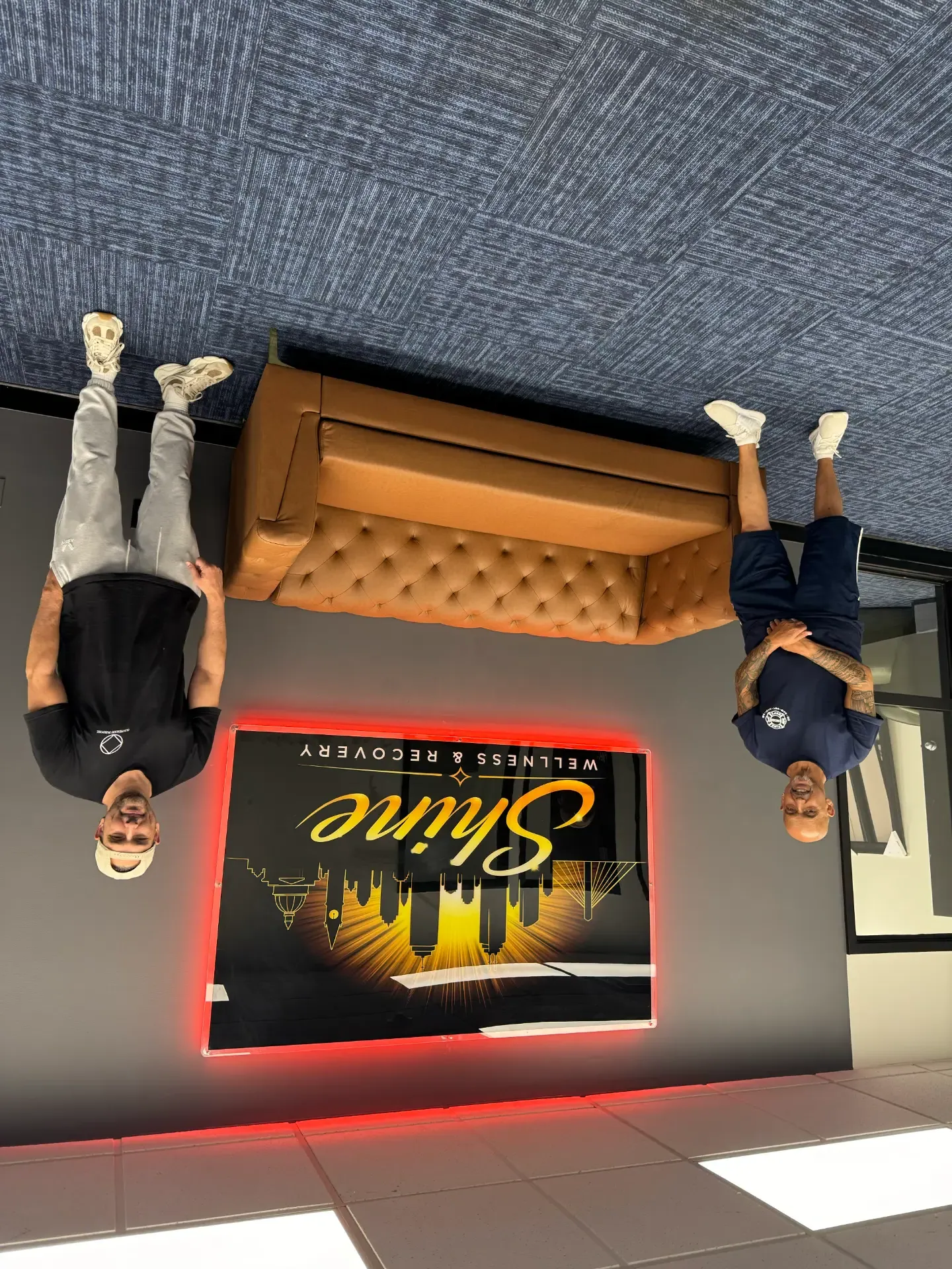 Two men standing in the Shine Wellness & Recovery lobby beside a tan leather sofa and an illuminated Shine Wellness & Recovery sign on the wall