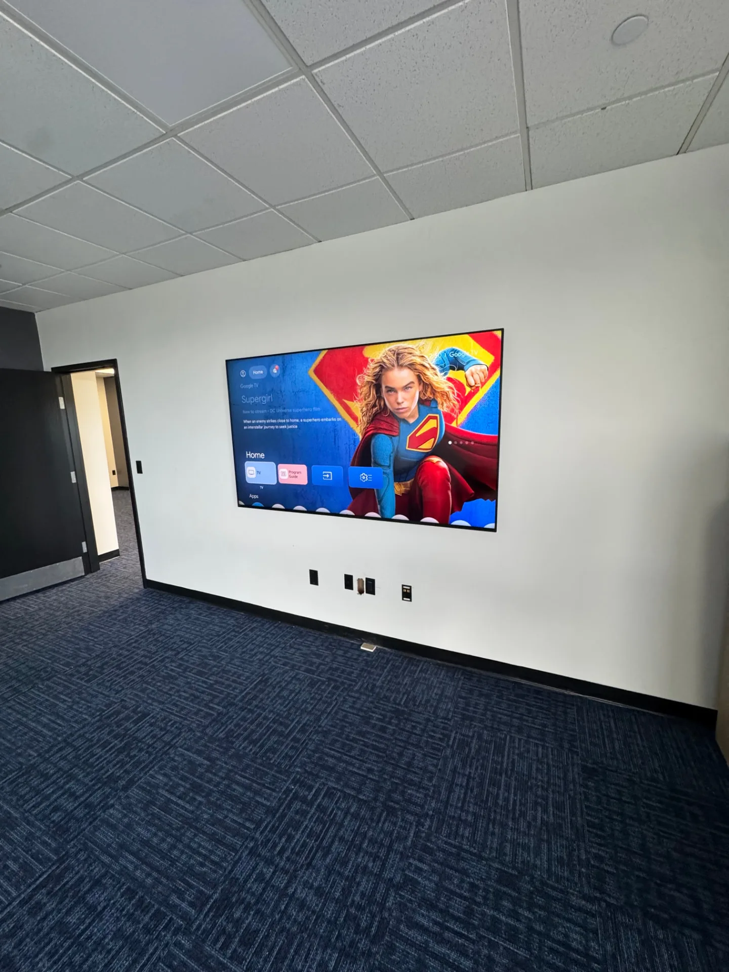 Group room at Shine Wellness & Recovery with a wall-mounted TV displaying the Google TV home screen, dark blue patterned carpet, and white walls