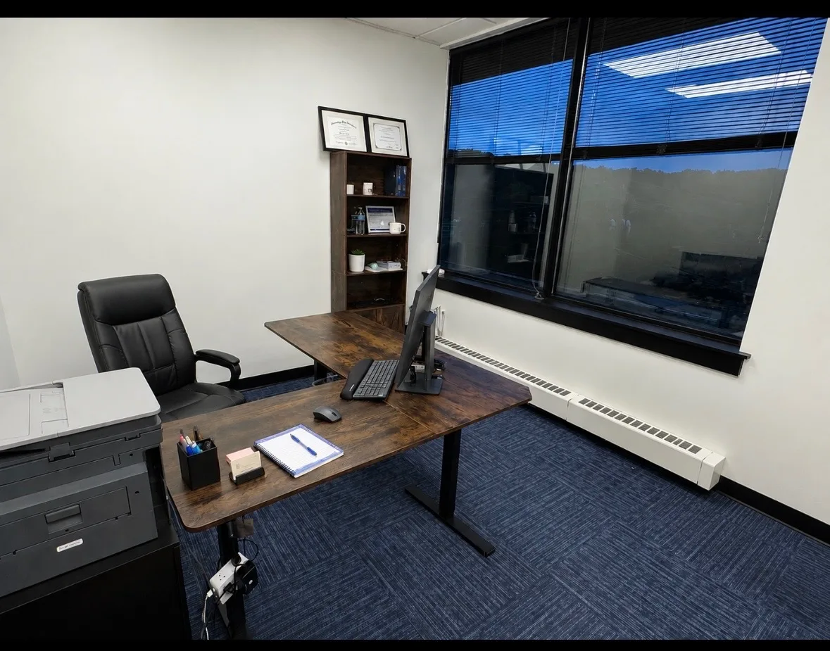 Furnished private office at Shine Wellness & Recovery with an L-shaped dark wood desk, black leather chair, bookshelf, and a window with black blinds