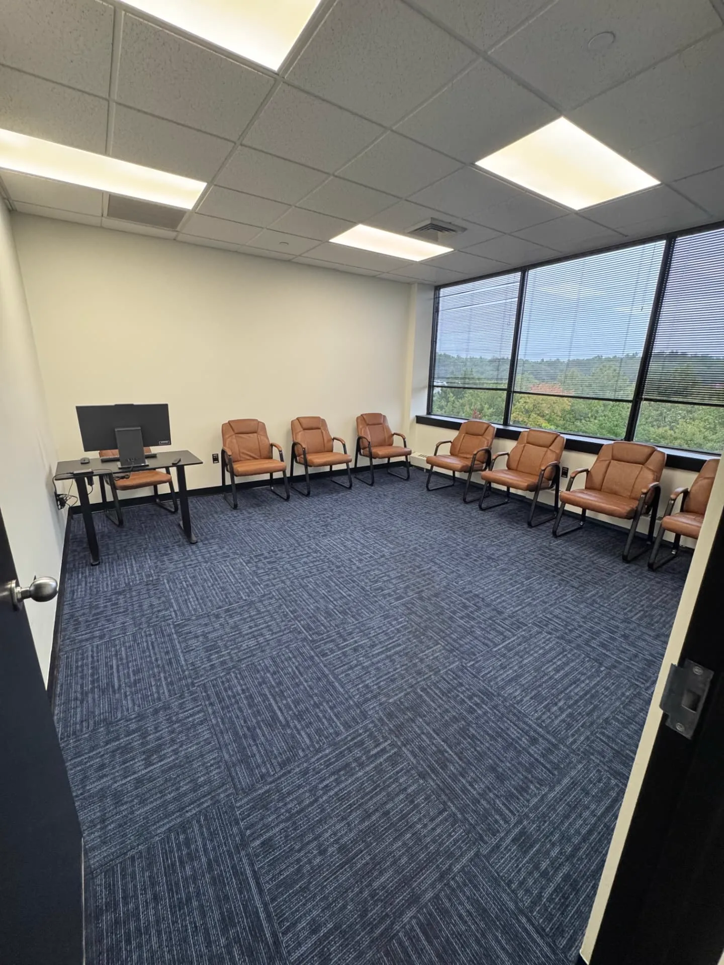 Small counseling room at Shine Wellness & Recovery with brown leather chairs along the window wall, a desk with a monitor, and blue patterned carpet