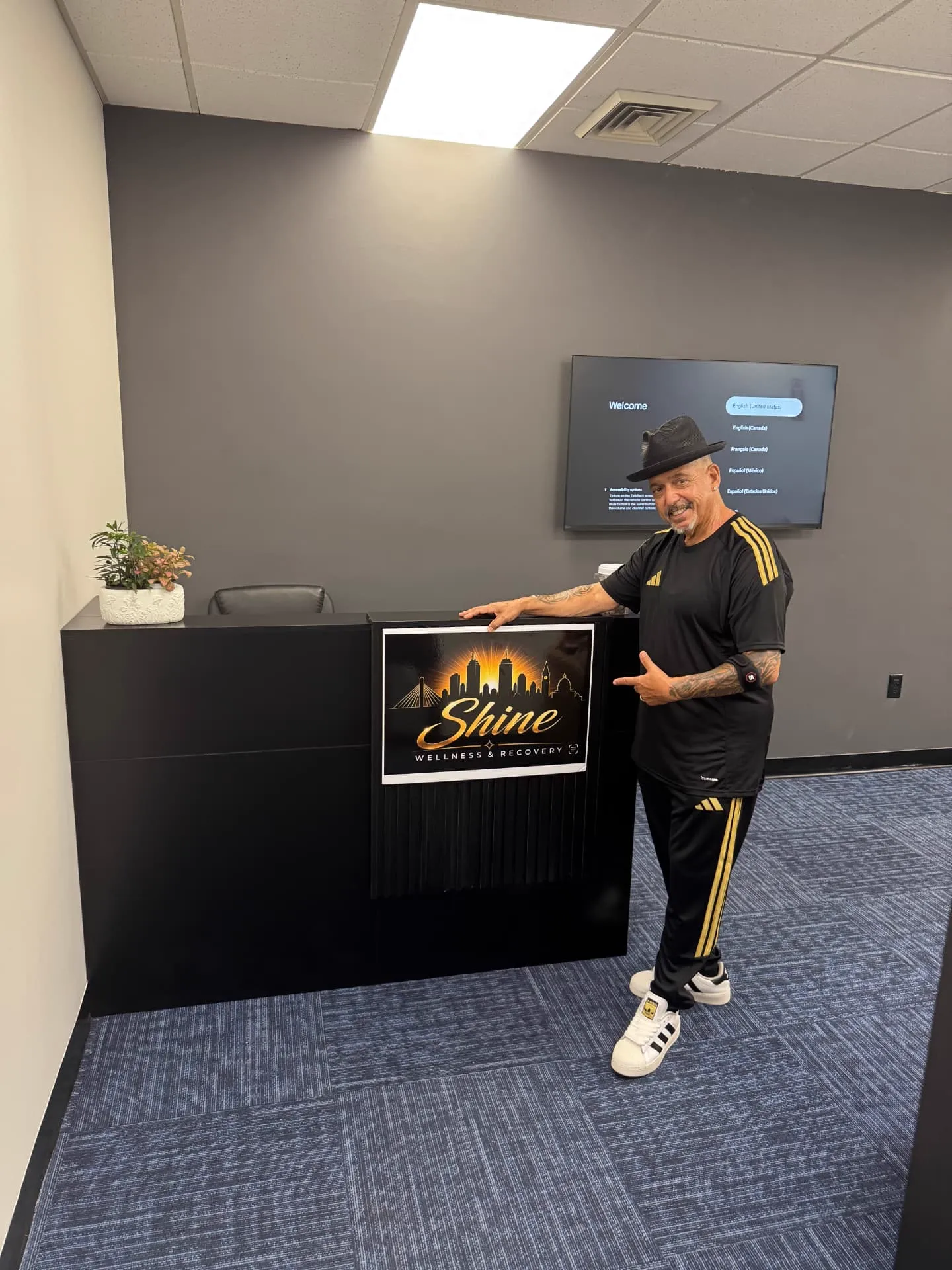 Reception area at Shine Wellness & Recovery with a black Shine-branded desk, wall-mounted TV showing a welcome screen, and blue patterned carpet